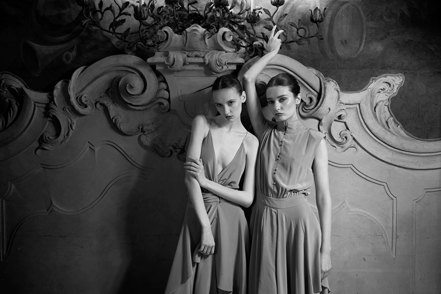 Two women in elegant dresses posing against an ornate wall.